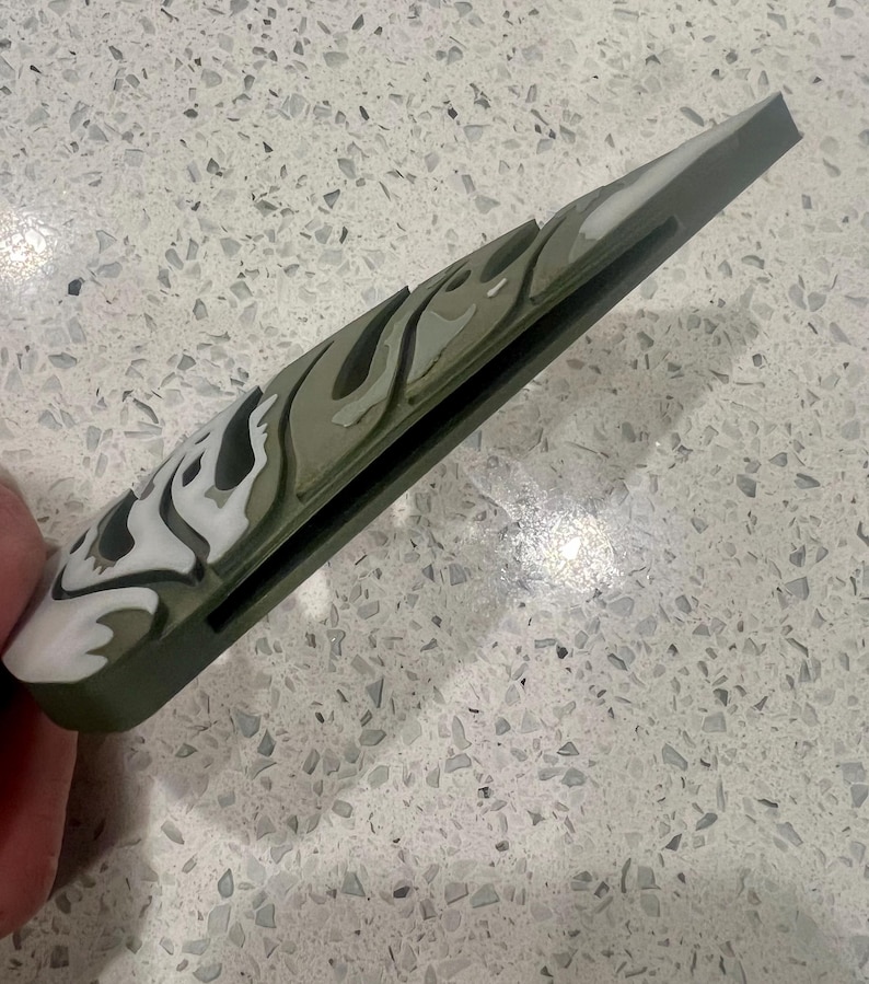 Puede incluir: Una funda de tel&eacute;fono de camuflaje verde y blanco con una ranura negra en el lateral.