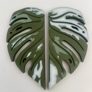 May include: Two green and white monstera leaf-shaped pieces of wall decor. The leaves are painted with a white and green camouflage pattern.
