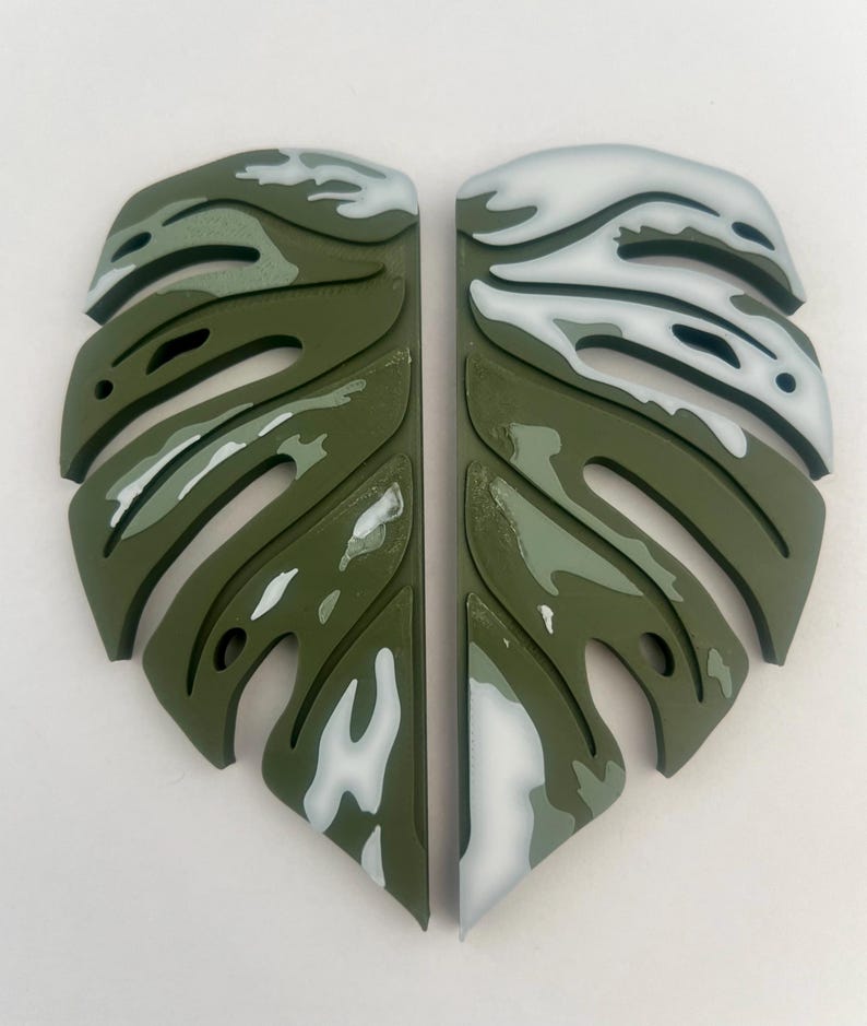 Puede incluir: Dos piezas de decoraci&oacute;n de pared en forma de hoja de monstera, verde y blanca. Las hojas est&aacute;n pintadas con un patr&oacute;n de camuflaje blanco y verde.
