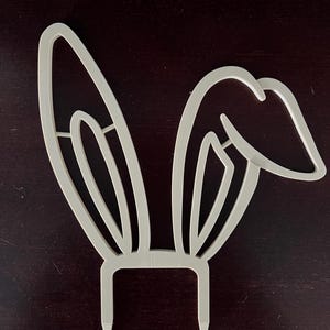 May include: White plastic bunny ear cake topper with two pointy prongs at the bottom.