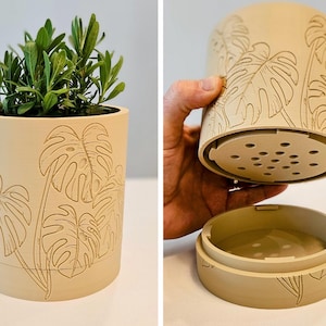 Puede incluir: Jardinera cilíndrica beige con un diseño detallado de hojas. La jardinera contiene una planta verde. La jardinera se muestra con su base separada y un inserto perforado. La jardinera mide aproximadamente 15 cm de alto y 15 cm de diámetro.