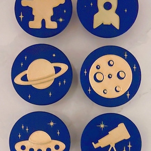May include: Six navy blue circular designs with gold-colored space-themed illustrations. The designs include an astronaut, rocket, Saturn, moon, UFO, and telescope, each surrounded by stars.