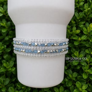 May include: A beaded wrap bracelet featuring alternating rows of clear glass beads, light blue faceted beads, and metallic silver beads. The bracelet is displayed on a white tumbler. The text SIP LUSH & CO TUMBLER ACCESSORY SHOP is visible.