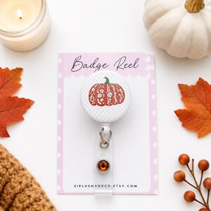 May include: A circular badge reel featuring a white and gray checkered pattern with a central orange pumpkin illustration decorated with white floral line art. The badge reel is attached to a white display card labeled Badge Reel.