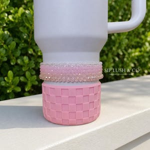 May include: A white tumbler with a pink checkered base and two bands of pink and clear beads. The text "SIP LUSH & CO" is printed on the side. The tumbler is on a white surface with green foliage in the background.
