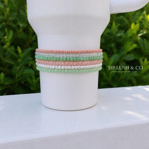 May include: A white tumbler featuring a stack of six beaded bracelets in alternating shades of peach, clear, light green, and white. The text SIP LUSH & CO TUMBLER ACCESSORY SHOP is printed in black on the right side of the white tumbler surface.