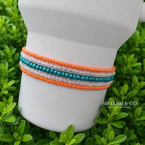 May include: A beaded bracelet featuring alternating rows of vibrant orange, clear, and teal beads. The bracelet is wrapped around a white cylindrical object. The text SIP LUSH & CO TUMBLER ACCESSORY SHOP is printed in the lower right corner of the image.