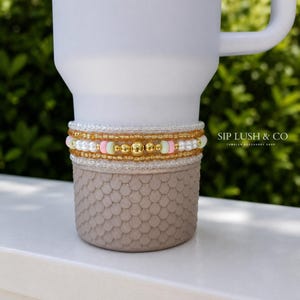 May include: A white tumbler with a beaded accessory. The accessory features gold, pink, and white beads. The bottom of the tumbler has a textured, scale-like design. The text "SIP LUSH & CO" is visible.