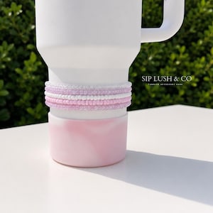 May include: A white tumbler with a pink and white base, adorned with a stack of beaded bracelets in shades of pink and white. The text "SIP LUSH & CO TUMBLER ACCESSORY SHOP" is visible on the right side of the image.