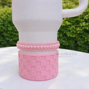 May include: A white tumbler with a pink beaded band and a pink checkered bottom. The tumbler has a handle and is set against a green background. The text "SIP LUSH & CO" is visible on the bottom.
