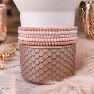 May include: Stack of beaded bracelets in shades of pink and white. The bracelets are displayed on a decorative, rose-gold container with a scale-like pattern. The image has a soft, feminine aesthetic.