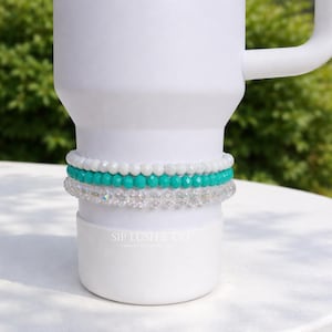 May include: A white tumbler with three beaded bracelets. The bracelets are white, turquoise, and clear crystal. The text "SIP LUSH & CO" is printed on the tumbler.