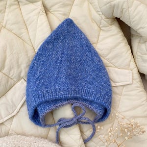 Könnte beinhalten: Eine blaue gestrickte Babyhaube mit spitzer Oberseite und Bindebändern. Die Haube ist aus weichem, strukturiertem Garn gefertigt. Der Hintergrund ist eine gesteppte cremefarbene Jacke.