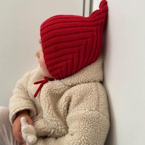 May include: A child wearing a bright red knitted hat with a distinctive pointed top. The hat has a ribbed texture. The child is also wearing a cream-coloured, fluffy jacket. A red tie is visible on the jacket.