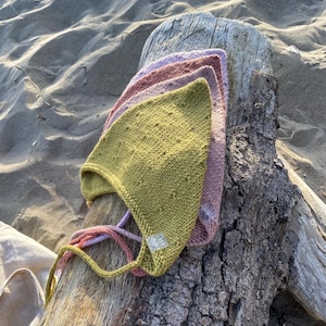 Op de afbeelding: Drie gebreide baby mutsjes in groen, roze en paarse tinten zijn gestapeld op een verweerde houten stam. De mutsjes hebben een textuur gebreid patroon en striksluitingen. De achtergrond toont zand en drijfhout.