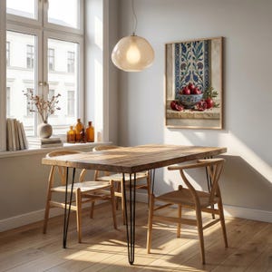 May include: A dining room scene featuring a wooden table with black hairpin legs and four wooden chairs. A framed still life painting of pomegranates hangs on the wall. A light fixture hangs above the table, and a window is visible in the background.