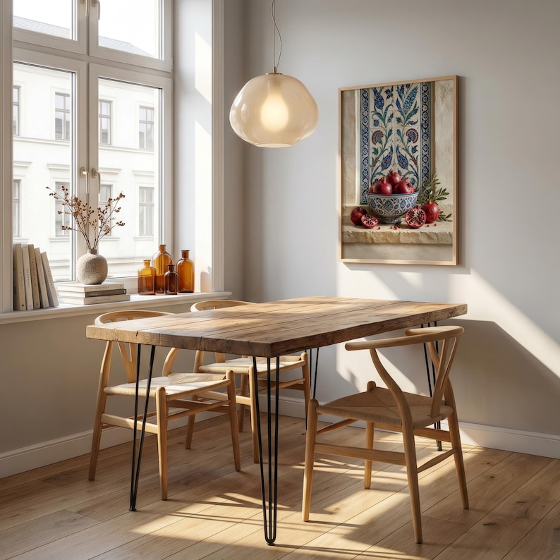 May include: A dining room scene featuring a wooden table with black hairpin legs and four wooden chairs. A framed still life painting of pomegranates hangs on the wall. A light fixture hangs above the table, and a window is visible in the background.