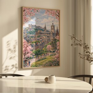 May include: A framed print of a cityscape, featuring a castle and buildings, framed by pink flowering trees. The artwork has a light wood frame and is hanging on a white wall. The scene is a watercolor painting with a soft, pastel color palette.