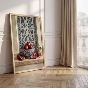 May include: A framed artwork featuring a still life with pomegranates. A decorative bowl filled with ripe pomegranates sits in front of a wall panel with blue and white floral patterns. The artwork is leaning against a white wall.