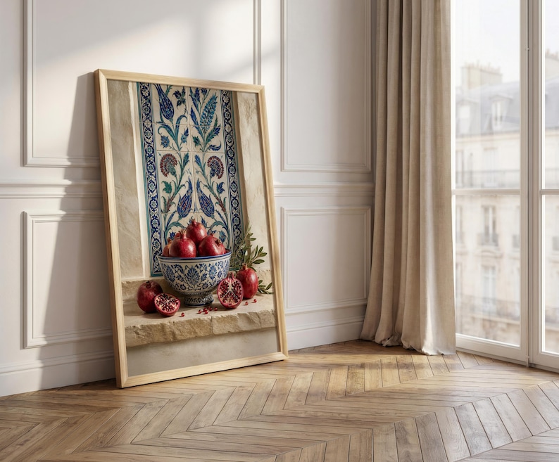 May include: A framed artwork featuring a still life with pomegranates. A decorative bowl filled with ripe pomegranates sits in front of a wall panel with blue and white floral patterns. The artwork is leaning against a white wall.