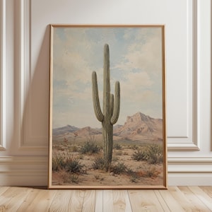 May include: A framed print of a desert landscape featuring a large saguaro cactus. The artwork has a vintage aesthetic, with a muted color palette of beige, green, and blue. Mountains are visible in the background under a cloudy sky.