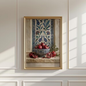 May include: A framed still life print featuring a bowl of pomegranates. The bowl is blue and white, filled with ripe red pomegranates. The background includes a decorative tile design and a stone ledge. The frame is gold.