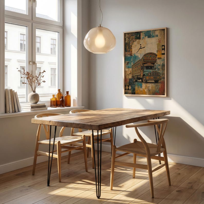 Op de afbeelding: Een rechthoekige houten eettafel met dunne zwarte metalen haarspeldpoten, vergezeld door drie lichtgekleurde houten stoelen met gevlochten zittingen. Een ingelijste kunstdruk van een versierde vrachtwagen met Arabische tekst hangt aan de muur.