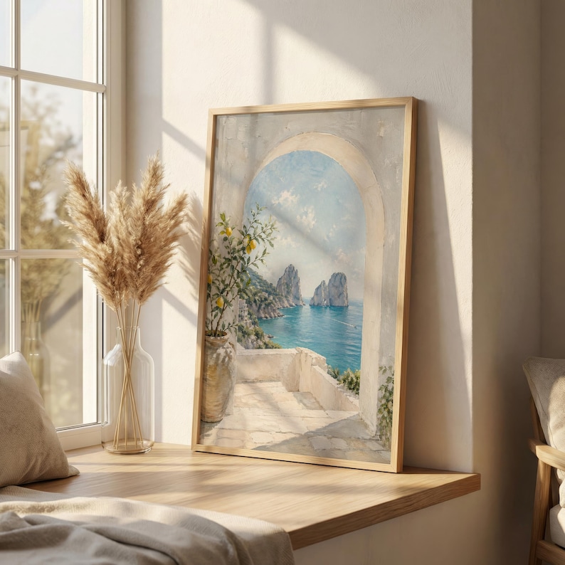 May include: A framed art print depicts a coastal scene with a stone archway overlooking the sea. The artwork features a vibrant blue ocean, rocky cliffs, and a lemon tree in a vase. The frame is light-colored, and the scene is illuminated by sunlight.