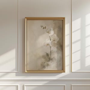 K&ouml;nnte beinhalten: Ein gerahmter Wandkunstdruck, der einen zarten wei&szlig;en Orchideenzweig vor einem weichen, neutralen Hintergrund mit texturierten, malerischen Pinselstrichen zeigt. Das Kunstwerk ist in einem d&uuml;nnen, goldfarbenen Metallrahmen eingefasst.