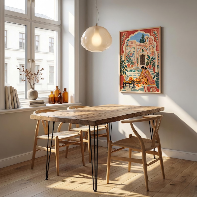 May include: A rectangular wooden dining table with black metal hairpin legs, surrounded by four light-colored wooden chairs with woven seats. A framed art print depicting a person in a courtyard hangs on the wall above the table.