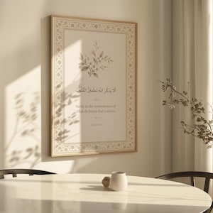 May include: A framed print featuring Arabic calligraphy and floral details. The text reads "Surely in the remembrance of Allah do hearts find comfort." The frame is light brown, and the print has a cream-coloured background.
