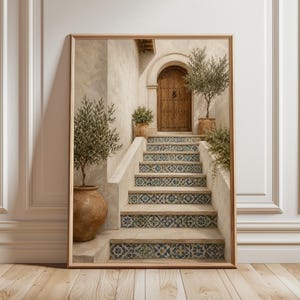 Könnte beinhalten: Gerahmter Kunstdruck, der eine mediterrane Szene darstellt. Eine Holztür befindet sich am oberen Ende einer Treppe mit blau-weiß gemusterten Fliesen. Olivenbäume in Terrakottatöpfen flankieren die Treppe. Das Kunstwerk befindet sich in einem Holzrahmen.