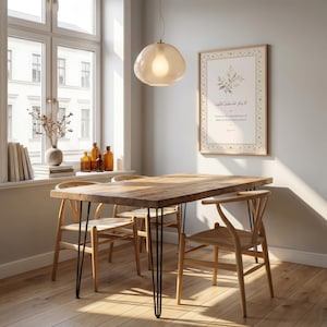 May include: A wooden dining table with black hairpin legs and four wooden chairs. A framed print with Arabic text hangs on the wall. A glass pendant light hangs above the table. The room has a window with a view.