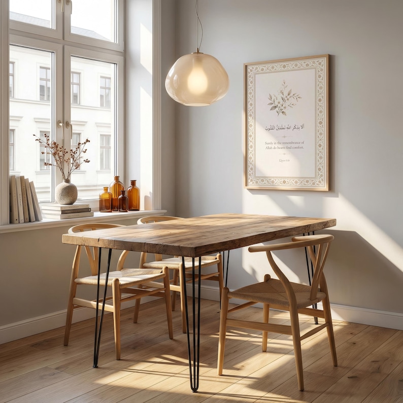 May include: A wooden dining table with black hairpin legs and four wooden chairs. A framed print with Arabic text hangs on the wall. A glass pendant light hangs above the table. The room has a window with a view.