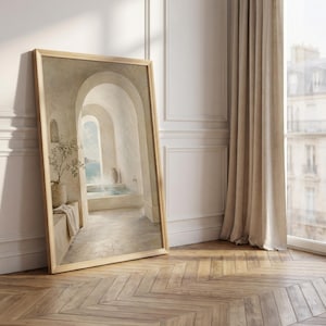 May include: A framed art print showing a tranquil interior scene with arched doorways and a pool. The artwork uses a neutral colour palette with beige and light blue tones. It is propped against a white wall and a wooden floor.