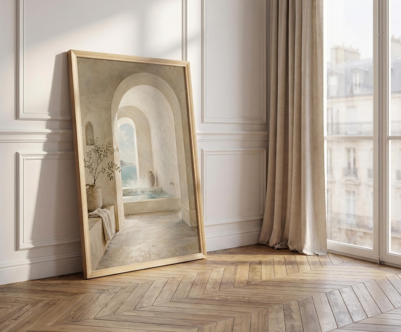 May include: A framed art print showing a tranquil interior scene with arched doorways and a pool. The artwork uses a neutral colour palette with beige and light blue tones. It is propped against a white wall and a wooden floor.
