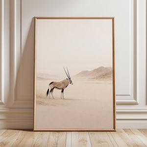 Op de afbeelding: Een rechthoekige wandkunstprint in een lichthouten lijst met een foto van een oryx die in een uitgestrekt, zanderig woestijnlandschap staat met verre bergen onder een bleke, wazige lucht. Het dier heeft lange, rechte, donkere hoorns en een beige vacht.