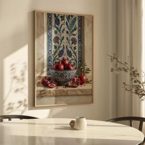 May include: A framed still life print featuring a bowl of pomegranates. The bowl is blue and white, filled with ripe red pomegranates. The background includes a decorative tile design with blue and green floral patterns. The print is in a wooden frame.