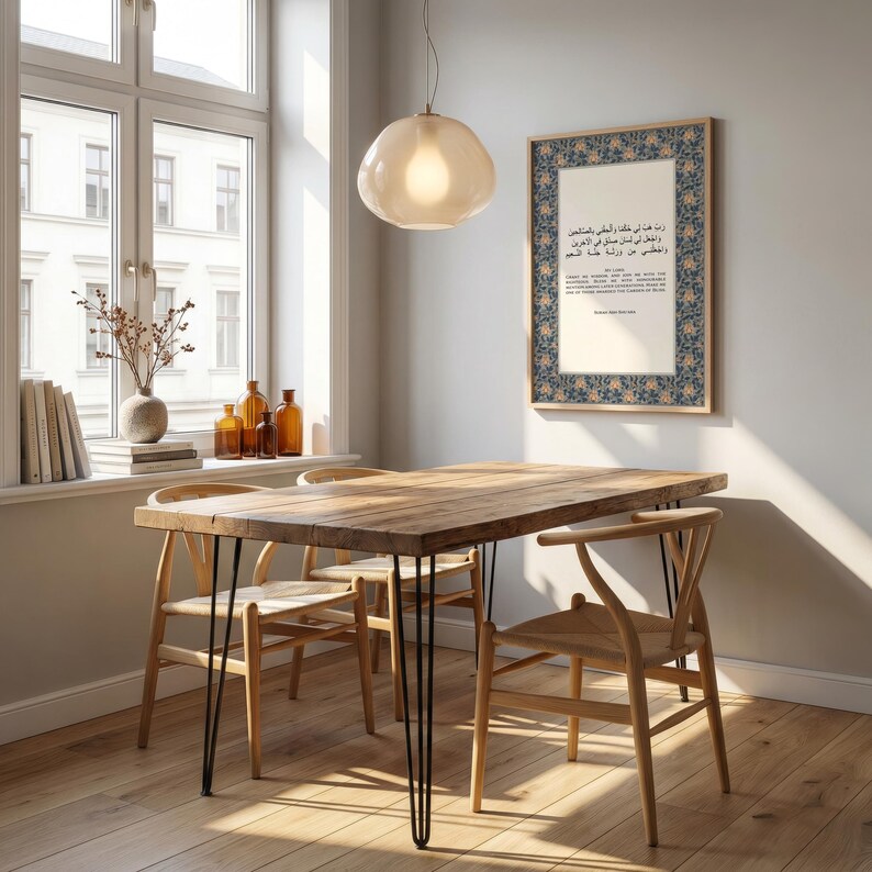 May include: A dining room scene featuring a wooden table with black hairpin legs and four wooden chairs. A framed print with Arabic text hangs on the wall. A glass pendant light hangs above the table. Decorative items are on the windowsill.