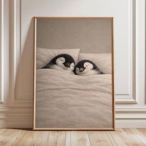 May include: A framed print showing two penguins asleep in a bed. The penguins are black and white with orange beaks, lying under a white duvet. The artwork is presented in a wooden frame.