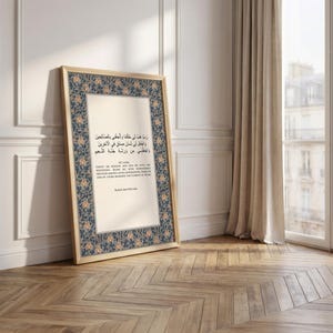 May include: A framed print with Arabic text and English text, featuring a floral border in blue and orange. The frame is light-colored, and the print leans against a white wall on a wooden floor.