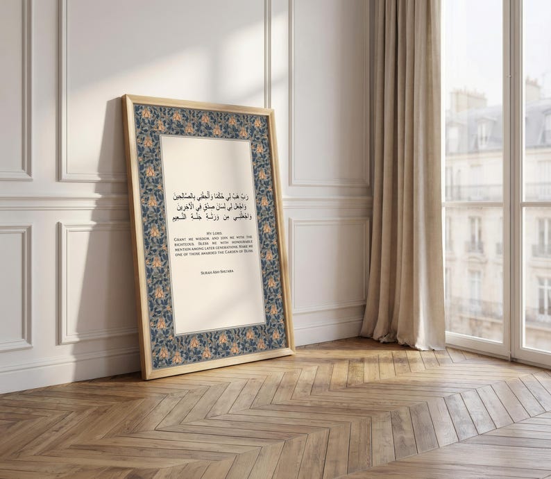 May include: A framed print with Arabic text and English text, featuring a floral border in blue and orange. The frame is light-colored, and the print leans against a white wall on a wooden floor.