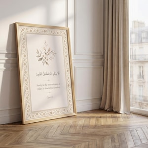 May include: A framed print with a light wood frame, leaning against a white wall. The print features Arabic calligraphy and English text: "Surely in the remembrance of Allah do hearts find comfort." A floral design accents the top. The room has a wooden floor.