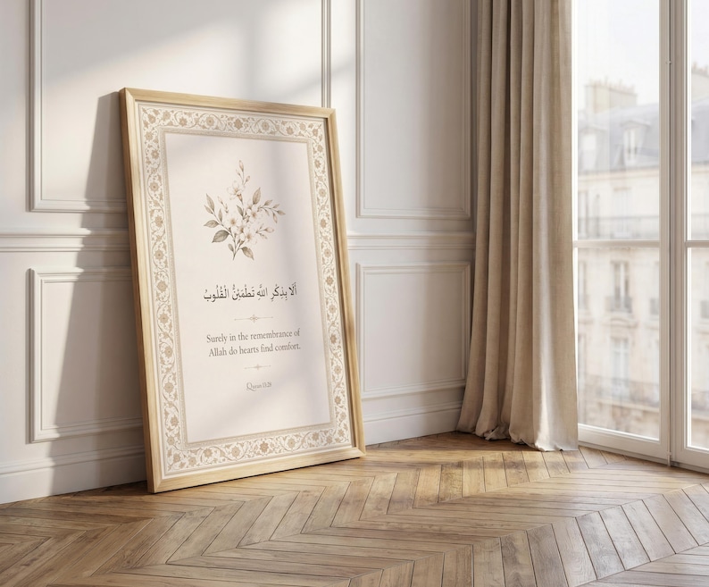 May include: A framed print with a light wood frame, leaning against a white wall. The print features Arabic calligraphy and English text: "Surely in the remembrance of Allah do hearts find comfort." A floral design accents the top. The room has a wooden floor.