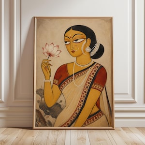 May include: A framed painting depicting a person with dark hair in a bun, wearing a red and cream sari with a patterned border. The figure is adorned with a nose ring, earrings, and a pearl necklace, holding a pink lotus flower in one hand.