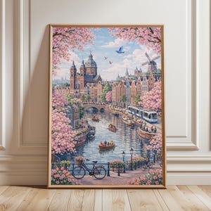 Könnte beinhalten: Ein gerahmter Druck von Amsterdam, der eine Kanalszene mit Booten, Gebäuden und einer Windmühle zeigt. Rosa Kirschblüten umrahmen das Bild, mit einem Fahrrad und Blumen im Vordergrund. Das Kunstwerk befindet sich in einem goldenen Rahmen.