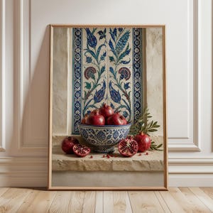 May include: A framed print featuring a still life of pomegranates in a decorative blue and white bowl. The bowl sits on a stone ledge in front of a wall with blue and white floral tiles. Several whole and halved pomegranates are visible.