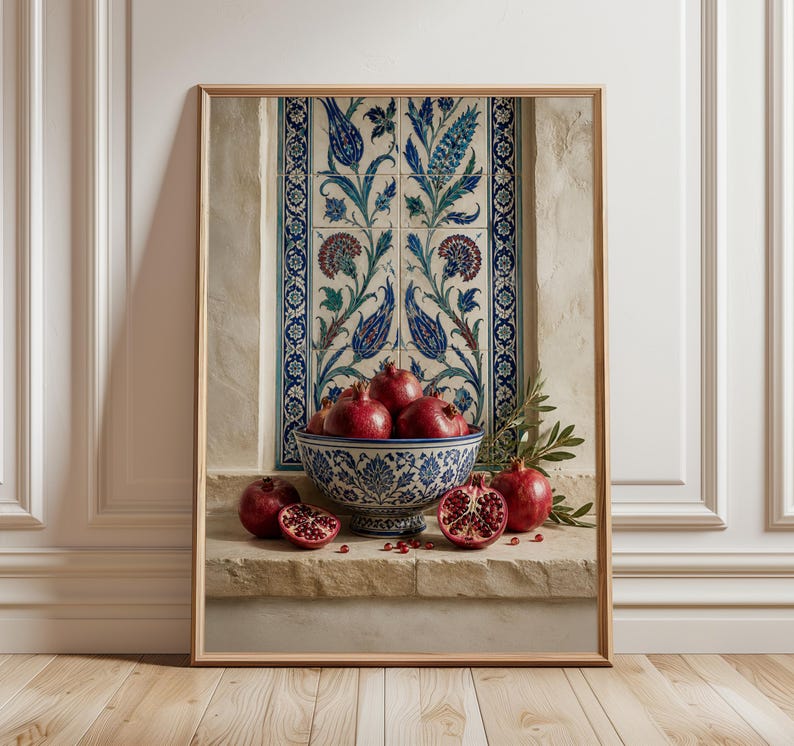 May include: A framed print featuring a still life of pomegranates in a decorative blue and white bowl. The bowl sits on a stone ledge in front of a wall with blue and white floral tiles. Several whole and halved pomegranates are visible.