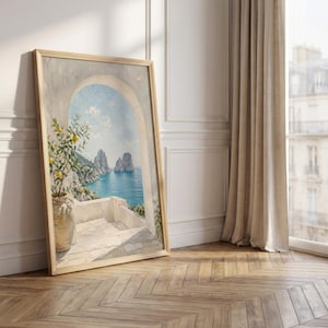 May include: A framed landscape painting depicts a coastal scene with a blue sea, rocky islands, and a white terrace with a lemon tree. The artwork is in a light wooden frame, leaning against a white wall and a wooden floor.