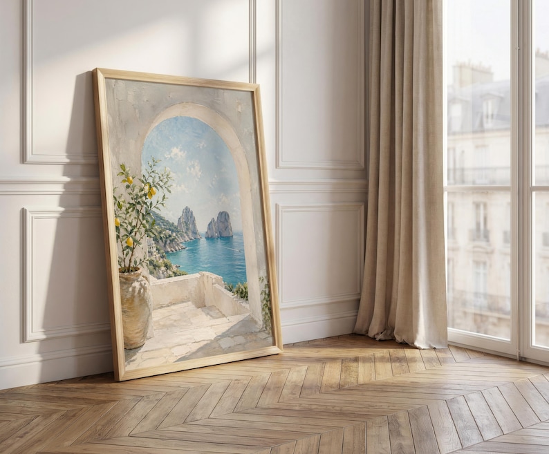 May include: A framed landscape painting depicts a coastal scene with a blue sea, rocky islands, and a white terrace with a lemon tree. The artwork is in a light wooden frame, leaning against a white wall and a wooden floor.